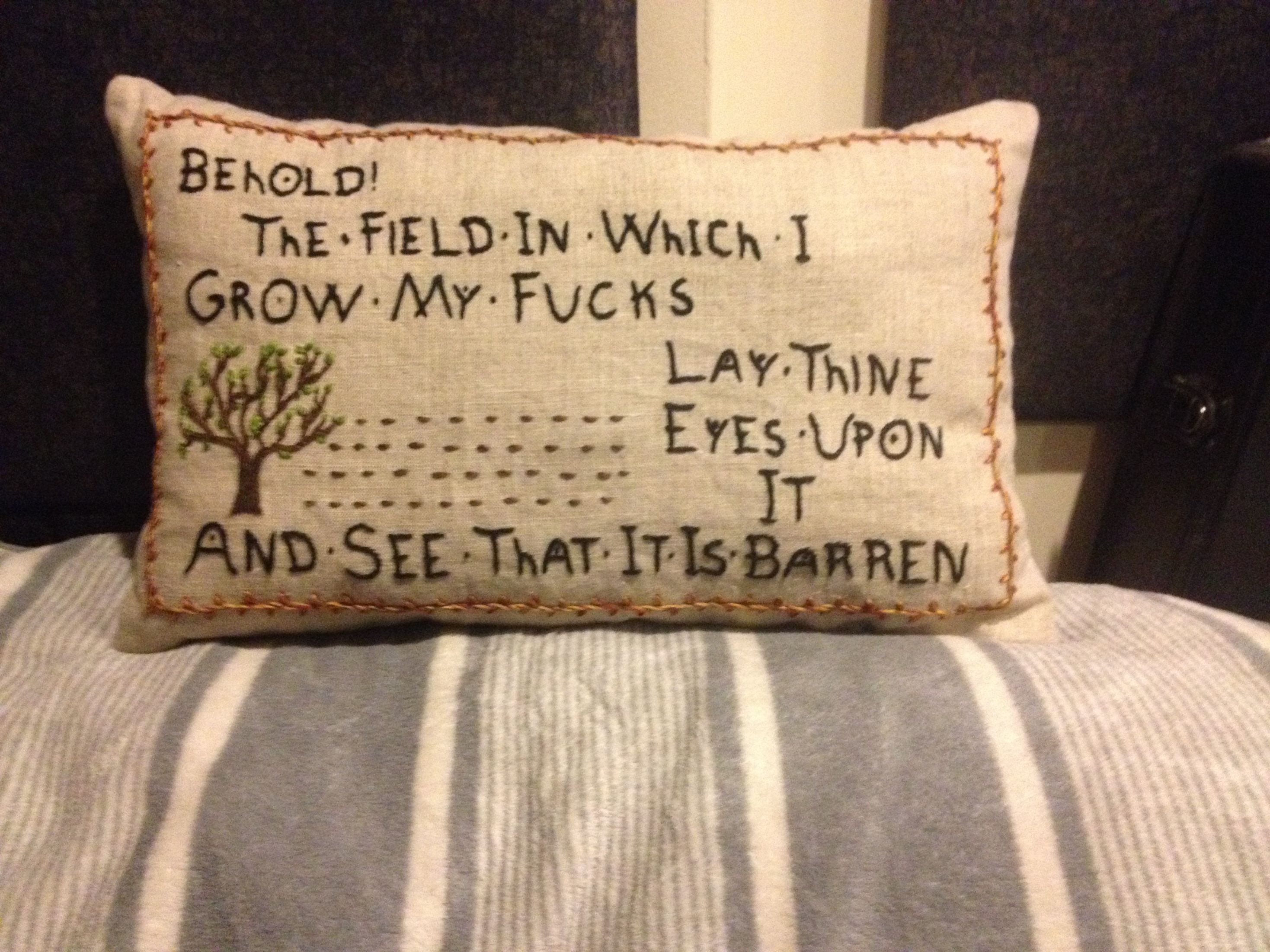 The embroidery of the text "Behold! The field on which I grow my fucks / Lay thine eyes upon/and see that it is barren." on a pillow, which rests on a grey-blanketed bed.